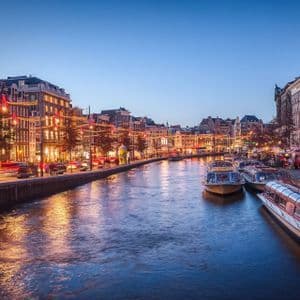 Un ampio canale cittadino al tramonto, con barche ormeggiate lungo la riva ed edifici illuminati che si riflettono sull'acqua.