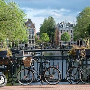Biciclette e una moto sono parcheggiate su un ponte cittadino che si affaccia su un canale costeggiato da edifici tradizionali e alberi verdi.