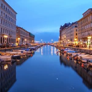Piccole imbarcazioni sono ormeggiate lungo un canale cittadino al crepuscolo, con edifici illuminati che si riflettono nell'acqua calma.