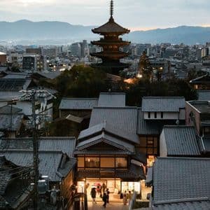 Una pagoda alta svetta su un panorama cittadino di tetti tradizionali al crepuscolo, con montagne lontane sotto un cielo nuvoloso.