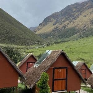 Una hilera de pequeñas cabañas rojizas con techos de paja y claraboyas en un frondoso valle verde de montaña.