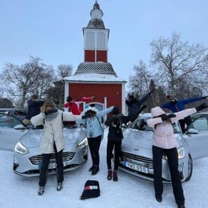 Eine WeRoad-Gruppe posiert im 'Dab'-Stil neben zwei Autos in einer verschneiten Landschaft, mit einem roten Gebäude im Hintergrund.