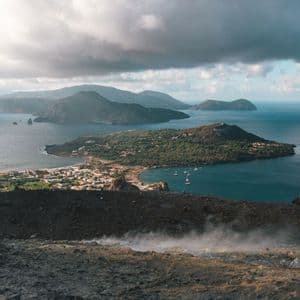 Une vue depuis un flanc de volcan fumant, dominant une ville côtière sur une île luxuriante, entourée par la mer et d'autres îles.