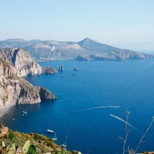 Vue imprenable sur une côte rocheuse et sauvage, baignée par une mer d'un bleu intense, avec des îles volcaniques à l'horizon et quelques bateaux.