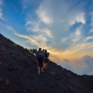 Un'escursione di gruppo WeRoad su un ripido pendio vulcanico scuro durante un tramonto colorato, con le nuvole visibili sotto.