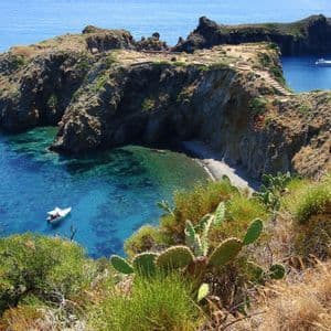 Blick von oben auf eine felsige Bucht mit klarem türkisfarbenem Wasser, einem kleinen Kieselstrand und mehreren Booten auf dem Meer.