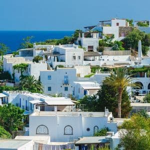 Un denso agglomerato di edifici imbiancati con vegetazione verde si erge su una collina che domina il mare blu intenso sotto un cielo limpido.