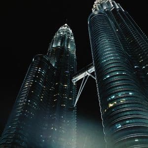 A low-angle view of two modern skyscrapers with a connecting sky bridge, illuminated against a dark night sky.