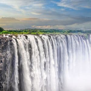 A massive waterfall rushes over a wide, rocky cliff edge, with green vegetation visible in the background under a blue and cloudy sky.