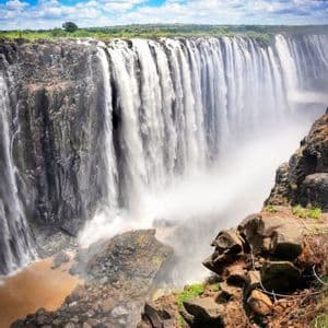 A massive waterfall cascades down a wide, sheer rock face into a misty gorge under a blue sky with white clouds.
