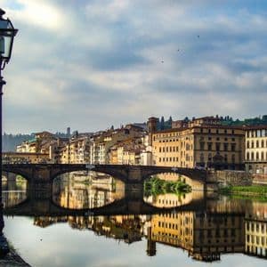 Un farol clásico se alza a orillas de un río, con un puente de piedra arqueado y edificios antiguos reflejados en las aguas tranquilas.