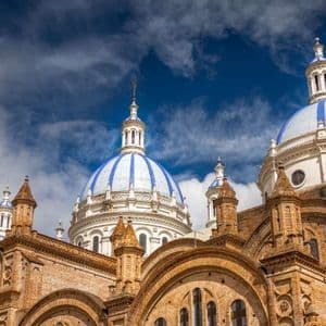A low-angle view of an ornate brick cathedral with large blue and white striped domes against a cloudy sky.