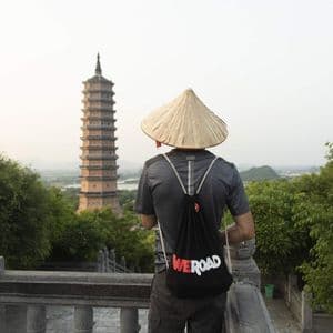 Un homme de dos portant un chapeau conique et un sac à dos WeRoad se tient sur un balcon en pierre dominant une haute pagode.