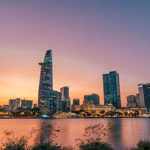 Uno skyline cittadino illuminato con moderni grattacieli che si riflettono in un fiume contro un cielo al tramonto viola e arancione.