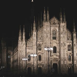 The intricate facade of a large Gothic cathedral with many spires, illuminated by lights against a dark night sky.