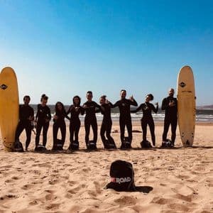 WeRoad Gruppenreise: Teilnehmer in Neoprenanzügen posieren mit Surfbrettern an einem sonnigen Strand mit dem Ozean im Hintergrund.