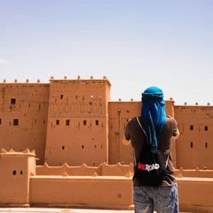 Una persona di spalle, con un foulard blu e uno zaino WeRoad, osserva una grande fortezza di mattoni di fango sotto un cielo azzurro.