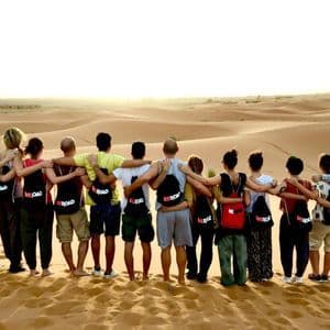 Un grupo de viaje de WeRoad, abrazados en una duna de arena, contemplando un vasto desierto al atardecer.
