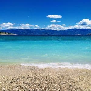 Una vista da una spiaggia di ciottoli attraverso acque turchesi verso una catena montuosa distante sotto un cielo azzurro parzialmente nuvoloso.