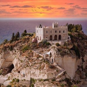 Un santuario in pietra su un'isola rocciosa si affaccia sull'oceano durante un tramonto vibrante, con una barca a vela in lontananza.