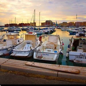 Molte barche sono ormeggiate in un porto tranquillo al tramonto, con edifici colorati sul lungomare sullo sfondo.