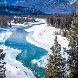Un fiume azzurro brillante si snoda attraverso una valle innevata circondata da pini e montagne distanti.