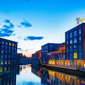 Edifici industriali in mattoni con finestre illuminate fiancheggiano un canale al tramonto, con le loro luci che si riflettono sulla superficie dell'acqua.