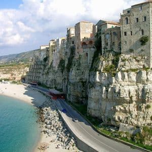 Edifici di una città costiera si ergono su una ripida scogliera, dominando una strada, una spiaggia sabbiosa e acqua turchese sotto un cielo azzurro.