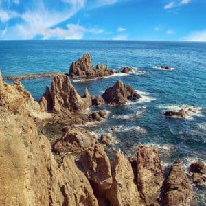 Formaciones rocosas dentadas emergen de un mar turquesa a lo largo de una escarpada costa de acantilados bajo un cielo azul con nubes tenues.