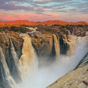 Una vasta cascata precipita da ripide scogliere rocciose in un canyon nebbioso, con montagne lontane illuminate dalla luce del tramonto.
