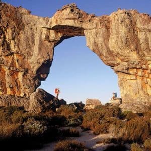 Due escursionisti con zaini si riposano sotto un enorme arco di roccia naturale in un paesaggio aspro, ricco di vegetazione secca, sotto un cielo limpido.