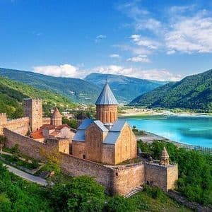 Un complexe castral fortifié avec une église se dresse sur une colline surplombant un lac turquoise et des montagnes verdoyantes.