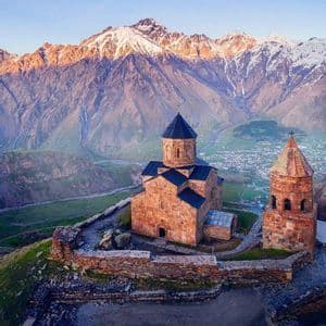 Une église historique en pierre et son clocher perchés sur une colline, avec une vaste chaîne de montagnes enneigées en arrière-plan au lever du soleil.