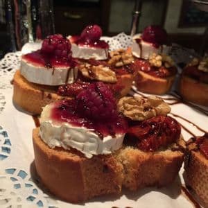 Crostini appetizers topped with goat cheese, raspberry jam, walnuts, and sun-dried tomatoes, drizzled with balsamic glaze.
