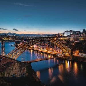 Un ponte ad arco metallico illuminato attraversa un ampio fiume al tramonto, con le luci della città che si riflettono sull'acqua.