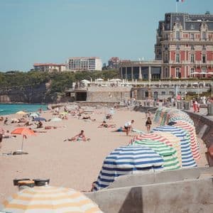 Menschen sonnen sich an einem Sandstrand voller bunter gestreifter Sonnenschirme, vor einem großen Gebäude mit französischer Flagge.