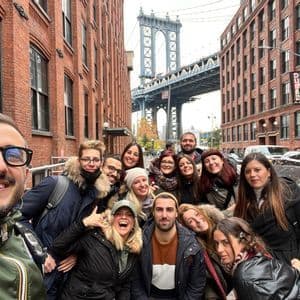 Un groupe WeRoad en voyage prenant un selfie souriant dans une rue urbaine, avec un grand pont visible entre des bâtiments en briques rouges.
