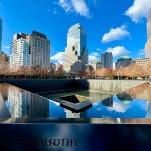 Una fuente conmemorativa cuadrada con nombres grabados en su borde, reflejando rascacielos y un cielo azul en el agua en un día soleado.