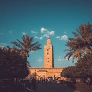 Un grand minaret orné se dresse derrière un bâtiment, encadré par des palmiers sur un ciel bleu avec des nuages épars.