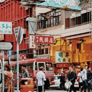 Persone e un minibus rosso su una trafficata strada cittadina piena di edifici colorati e grandi insegne con caratteri cinesi.