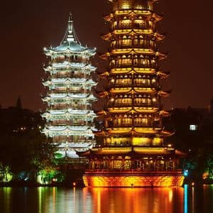Due alte pagode, una illuminata con luce dorata e una con luce bianca, che si riflettono su un lago calmo di notte.