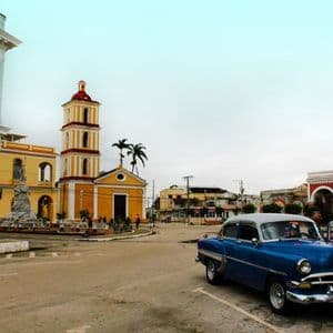 Une voiture bleue ancienne garée sur une place de ville devant une église jaune et des bâtiments coloniaux colorés.