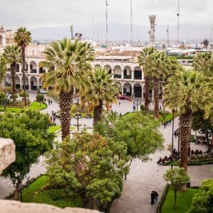An elevated view of a bustling city plaza filled with palm trees, green spaces, and people walking among historic buildings under a cloudy sky.