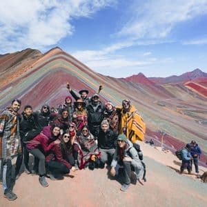 Un groupe WeRoad pose pour une photo sur le flanc d'une montagne avec une grande montagne rayée et colorée en arrière-plan sous un ciel bleu.