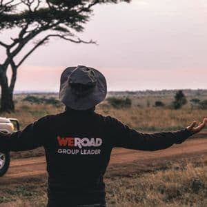 Ein WeRoad-Gruppenleiter steht, von hinten gesehen, mit ausgestreckten Armen in einer Savannenlandschaft neben einem Safari-Fahrzeug bei Sonnenuntergang.