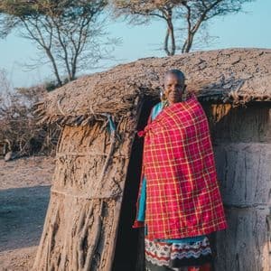 Una mujer mayor, vistiendo una manta roja a cuadros y atuendo tradicional, se para en la entrada de una cabaña de barro.