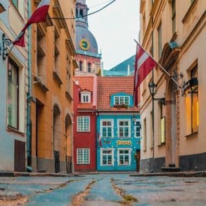 A low-angle view of a narrow cobblestone street leading to colorful buildings, with two Latvian flags hanging and a clock tower in the background.