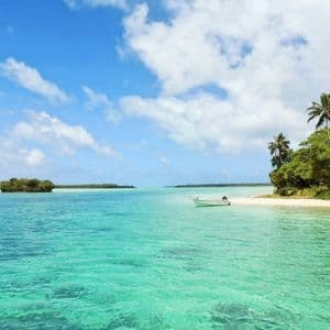 Una piccola barca è ormeggiata sulla spiaggia sabbiosa di un'isola tropicale con acqua cristallina e turchese sotto un cielo parzialmente nuvoloso.