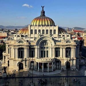 Un edificio neoclásico ornamentado con una cúpula de azulejos coloridos, visto desde arriba, con una plaza de la ciudad y montañas al fondo.