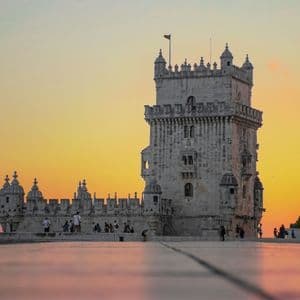 Una fortezza con torre in pietra ornata si erge sul lungomare al tramonto, vista da un'angolazione bassa su una piazza pavimentata.